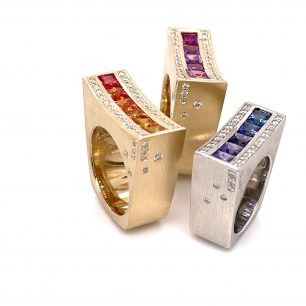 Three gold rings features gemstones in various colors such as red, violet and blue. The ring positioned upright set on white background.
